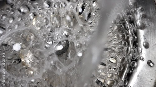 Water with bubbles of soap flows into the drain hole in the metal sink, macro view. Mechanically adjustable drain plug closeup. White foam with bubbles of cleaner in a washbasin. photo