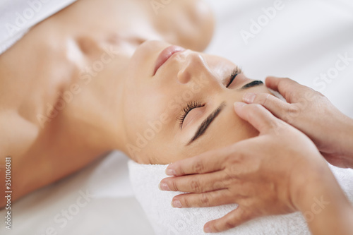 Close-up image of relaxed young woman getting rejuvenating face massage in beauty salon