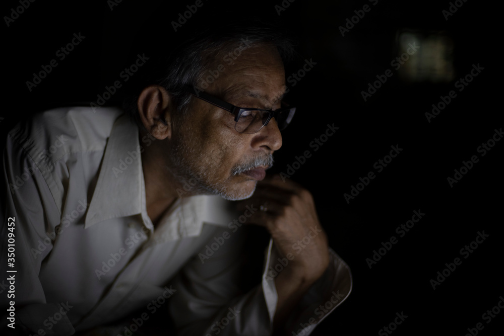 An Indian old man is watching laptop screen while video chatting leaning on his bed in the darkness. Indian lifestyle, home isolation and lock down