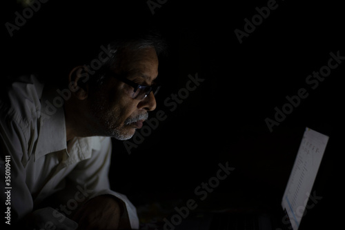 An Indian old man is watching laptop screen while video chatting leaning on his bed in the darkness. Indian lifestyle, home isolation and lock down