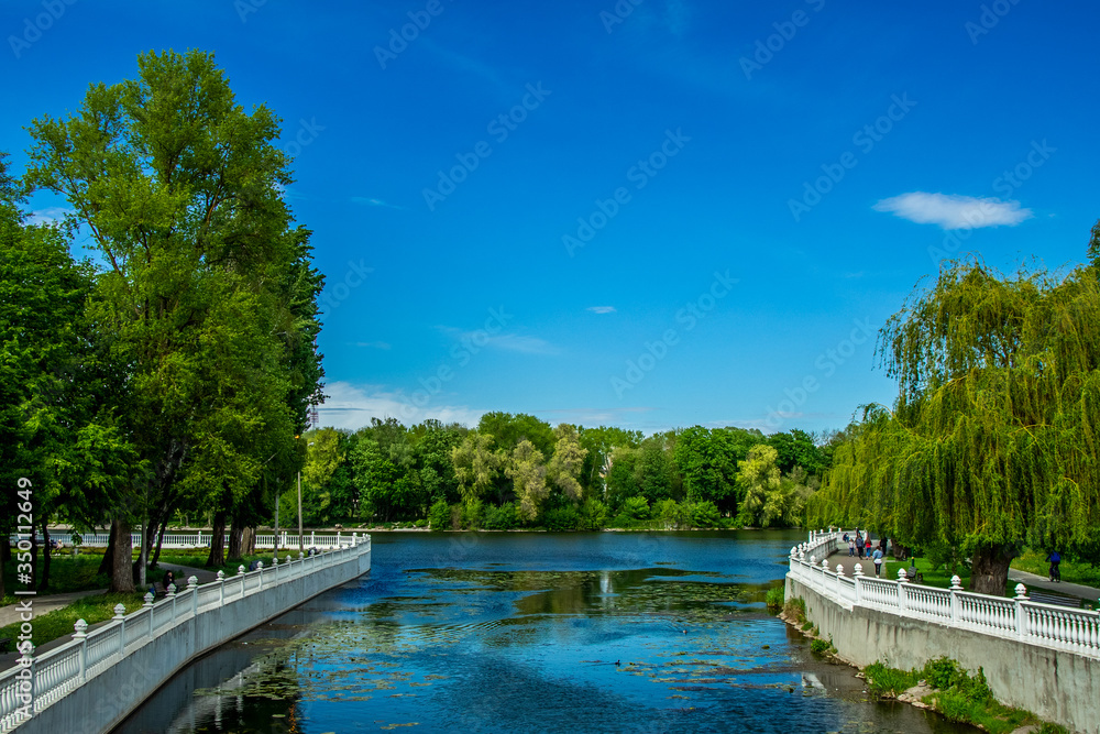Fototapeta premium summer day time in June city waterfront walking site district marble fence along river and vivid green trees foliage and blue sky background idyllic landscape scenic