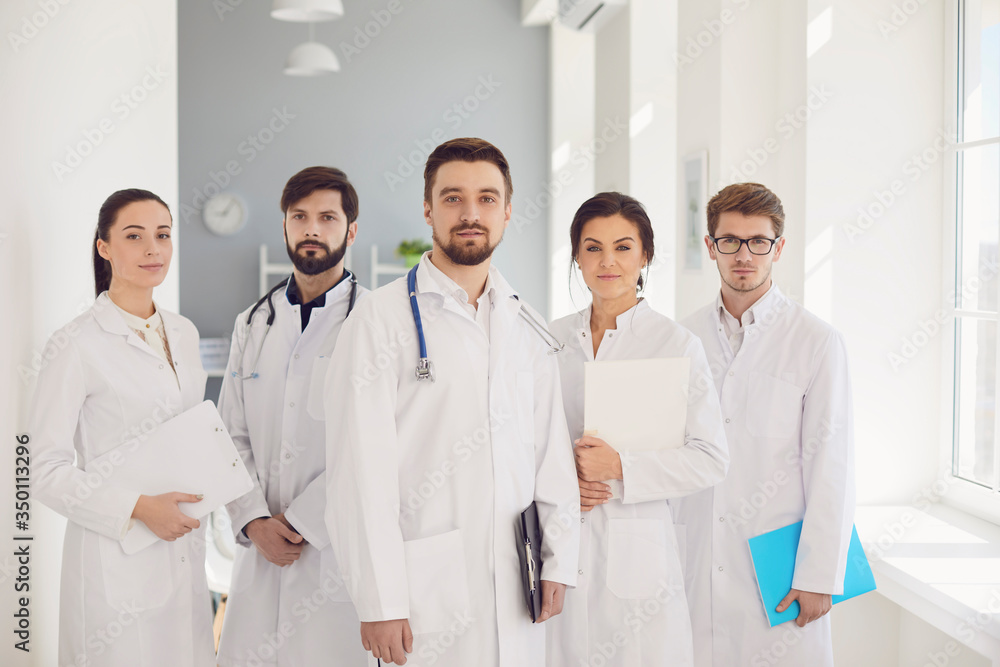 A group of confident practicing doctors in white coats are smiling against the backdrop of the clinic. A group of confident practicing doctors in white coats are smiling against the backdrop of the clinic.