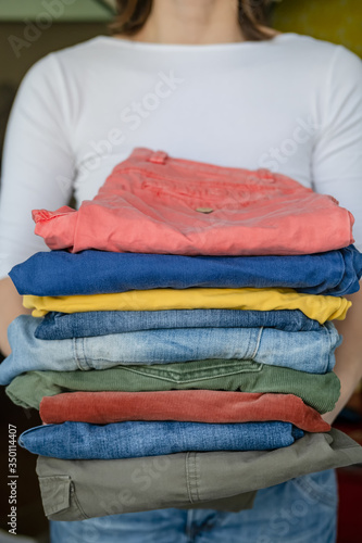 Woman holds neatly folded trousers and jeans in bright colors, after washing, in the laundry room.