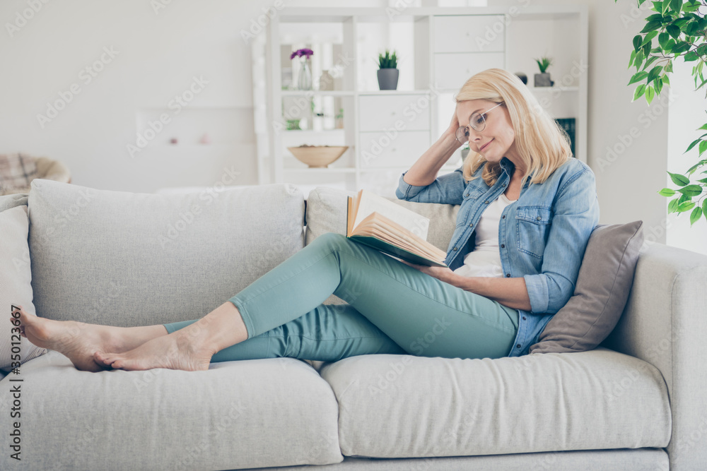 Profile photo of attractive aged mature homey lady sitting comfy sofa couch  barefoot reading interesting historic novel book stay home quarantine time  weekend living room indoors Stock Photo