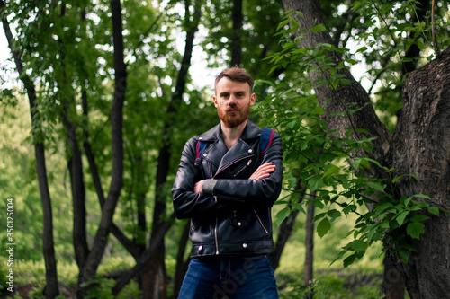A bearded man stands in the forest in a leather jacket with his arms crossed.