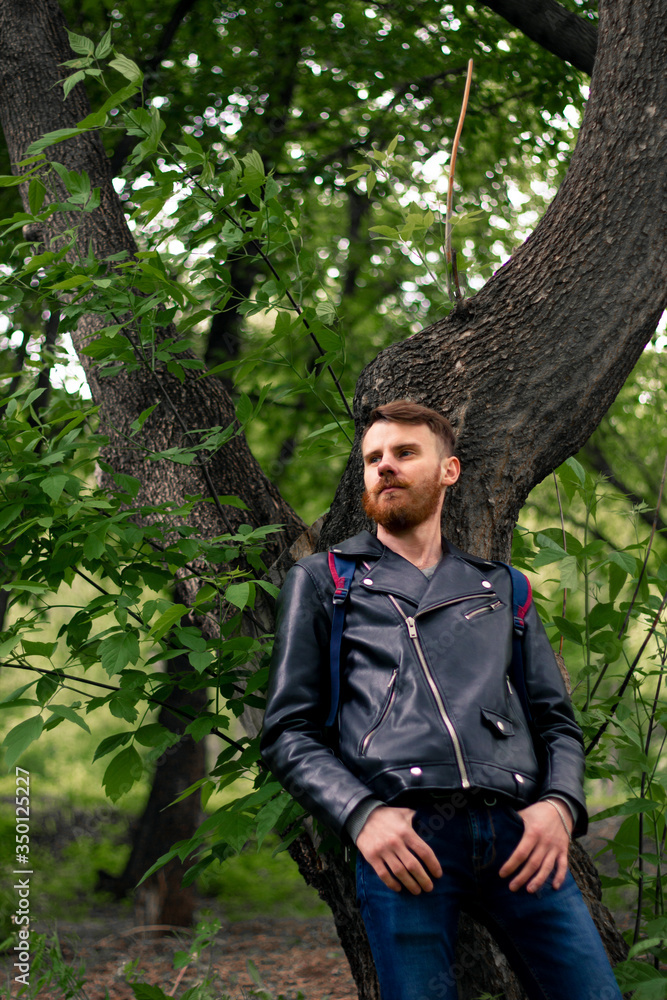 a man in a black leather jacket looks thoughtfully away, leaning his back against a tree. a man in a black leather jacket looks thoughtfully away, leaning his back against a tree.
