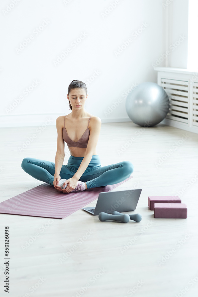 Minimal full length portrait of fit young woman warming up and stretching while watching workout video via laptop at home, copy space Minimal full length portrait of fit young woman warming up and stretching while watching workout video via laptop at home, copy space