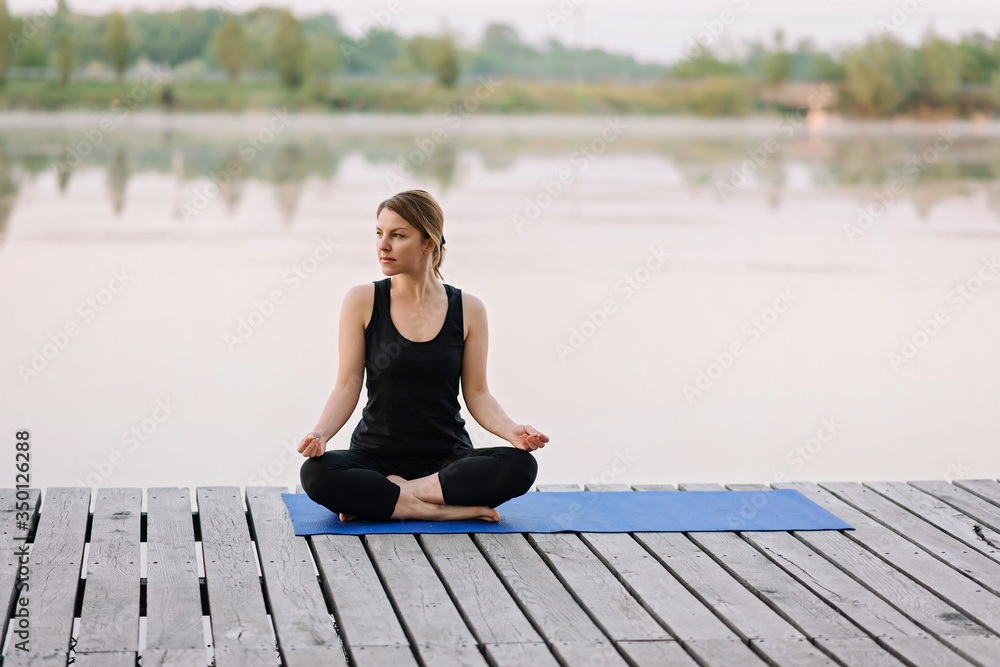 A 36-year-old young Caucasian woman practices yoga in the lotus position outdoors near the river on a wooden pier in the morning. Healthy lifestyle concept. Morning meditation, relaxation.