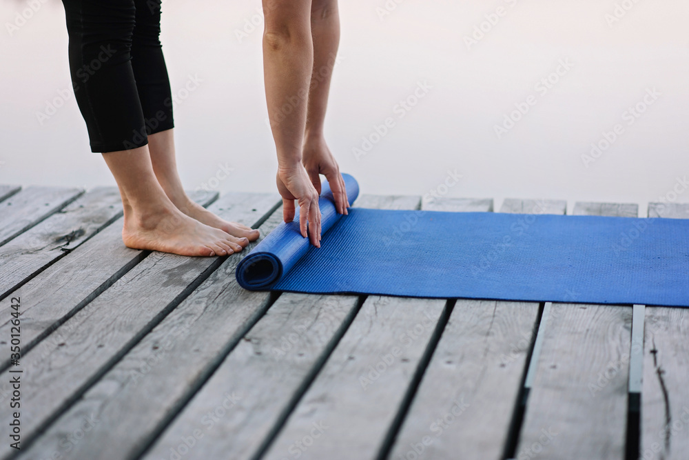 woman twists yoga mat outdoors near the river on a wooden pier in the morning. Healthy lifestyle concept. Morning meditation, relaxation.