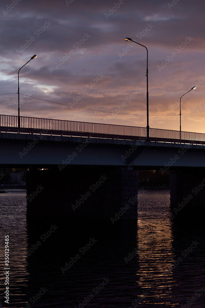 Wonderful evening dusk with sunset sky, clouds, golden sunbeam over black bridge over river with square frame and lantern light, minimalist landscape.