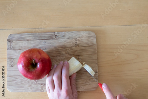 Kind bereitet sich gesundes Frühstück oder Snack aus Obst wie Apfel und Banane zu für eine gesunde Ernährung und einen gesunden Start in den Tag mit Vitaminen auf einem Bambusbrettchen in der Küche