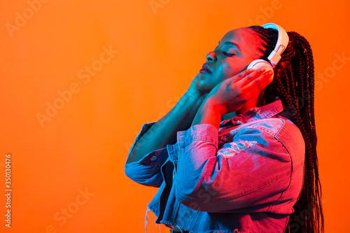 Close up beauty fashion portrait of an attractive young african woman wearing dreadlocks standing over orange background, listening to music with headphones. Neon light and copy space
