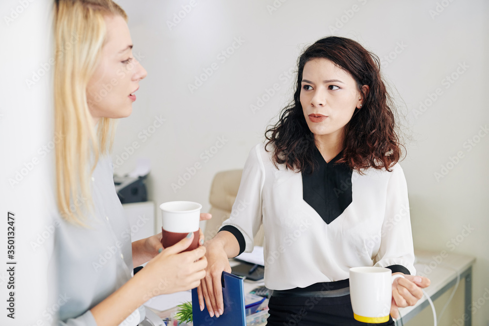 Serious young mixed-race businesswoman drinking cup of coffee and discussing project with colleague Serious young mixed-race businesswoman drinking cup of coffee and discussing project with colleague