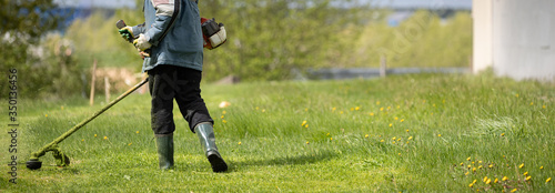A view from the back of a man with a lawn mower. Worker in protective clothing mows grass on the lawn with a trimmer. Wide view