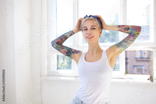 Sunny shot of young attractive tattooed blonde lady keeping raised hands on her head while looking at camera with pleasant smile  standing in front of big window