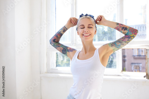 Pleasant looking young lovely tattooed blonde woman smiling nicely with closed eyes while stretching raised hands, posing over window on sunny warm day