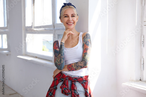 Canvas-taulu Cheerful young tattooed attractive female in headband and casual wear touching h