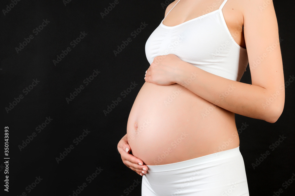 Close up of pregnant woman touching her abdomen at black background. Future mother is wearing white underwear. Expecting of a baby. Copy space Close up of pregnant woman touching her abdomen at black background. Future mother is wearing white underwear. Expecting of a baby. Copy space