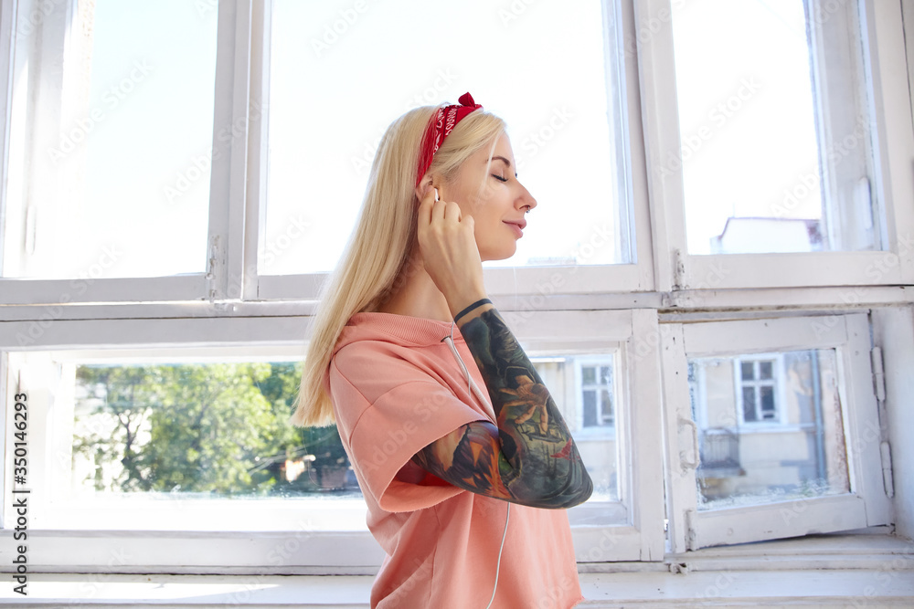 Side view of young tattooed blonde long haired woman inserting earpiece in her ear while walking over big window, keeping her eyes closed and smiling pleasantly