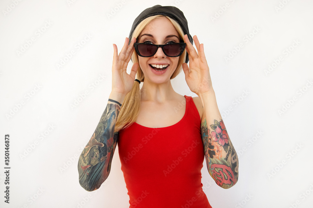 Cheerful young pretty tattooed blonde lady in elegant clothes keeping raised hand on her eyewear while looking gladly at camera with wide smile, isolated over white background Cheerful young pretty tattooed blonde lady in elegant clothes keeping raised hand on her eyewear while looking gladly at camera with wide smile, isolated over white background