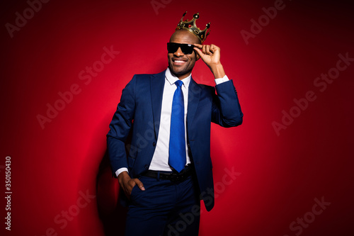 Photo of handsome selfish cool dark skin business guy bossy person golden diadem on head looking flirty eyes wear sun specs formalwear suit tuxedo isolated burgundy red background photo