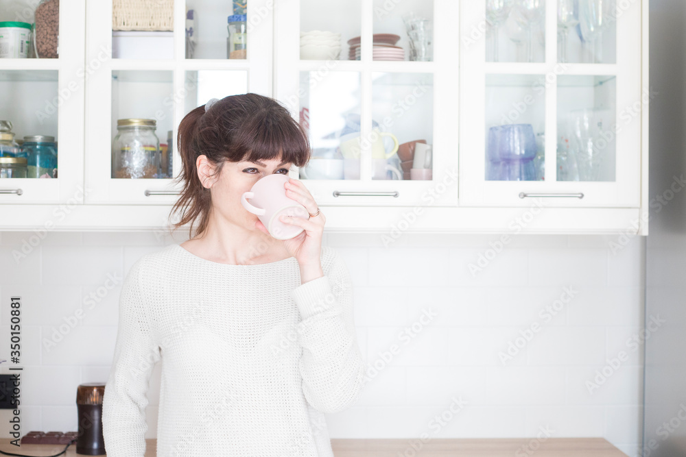 Pretty caucasian woman, with bangs and ponytail, drinking a cup of coffee in the morning in a white, bright lit, kitchen. Relaxed and positive atmosphere. Pretty caucasian woman, with bangs and ponytail, drinking a cup of coffee in the morning in a white, bright lit, kitchen. Relaxed and positive atmosphere.