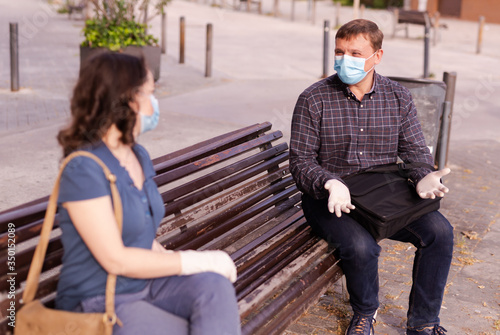 Emotional couple in protective medical masks against coronavirus communicate sittig on bench photo