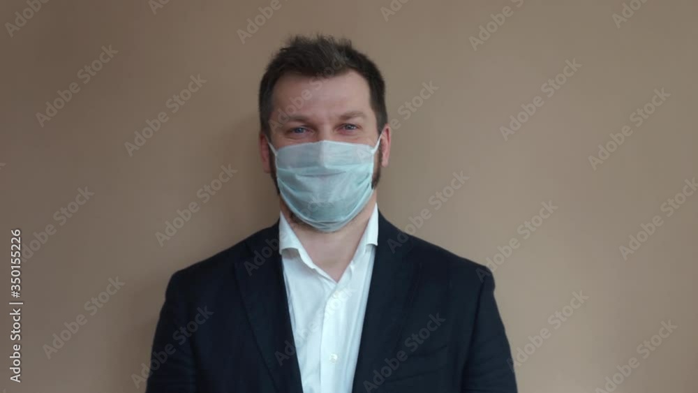 Handsome man in a business suit, a jacket and a white shirt puts on a protective medical mask and looks at the camera on a beige background. Natural light, soft focus. Handsome man in a business suit, a jacket and a white shirt puts on a protective medical mask and looks at the camera on a beige background. Natural light, soft focus.
