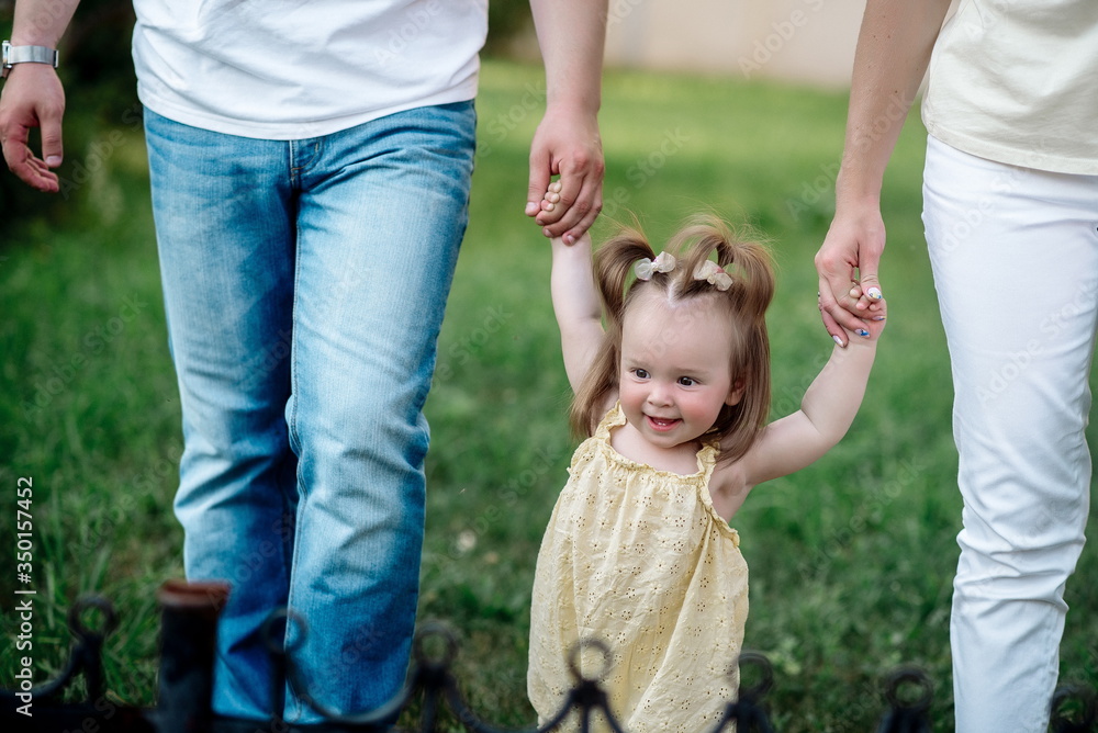 Fototapeta premium Dad and mom lead their funny one-year-old daughter in the park in summer 
