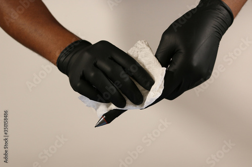 Pair of hands wearing black latex or vinyl glove wiping down desk equipment. Hands Wipe tablet pencil pen stylus down with paper towel and disinfectant  photo