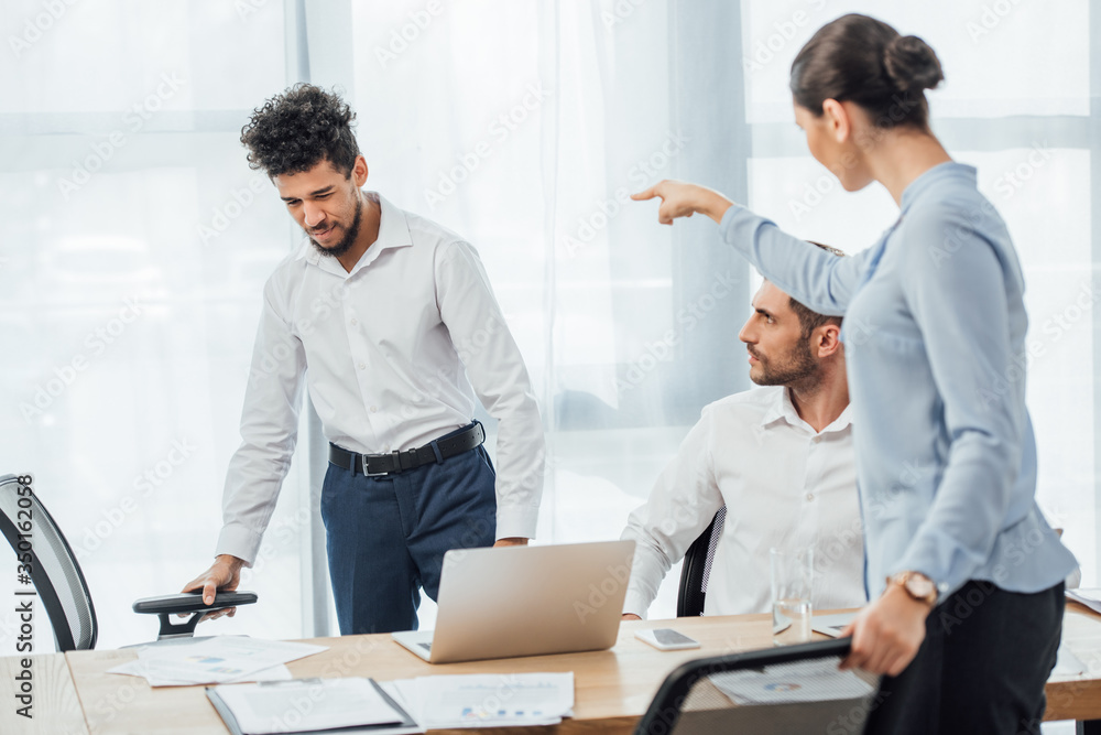 Selective focus of businesswoman pointing with finger at sad african american colleague in office Selective focus of businesswoman pointing with finger at sad african american colleague in office
