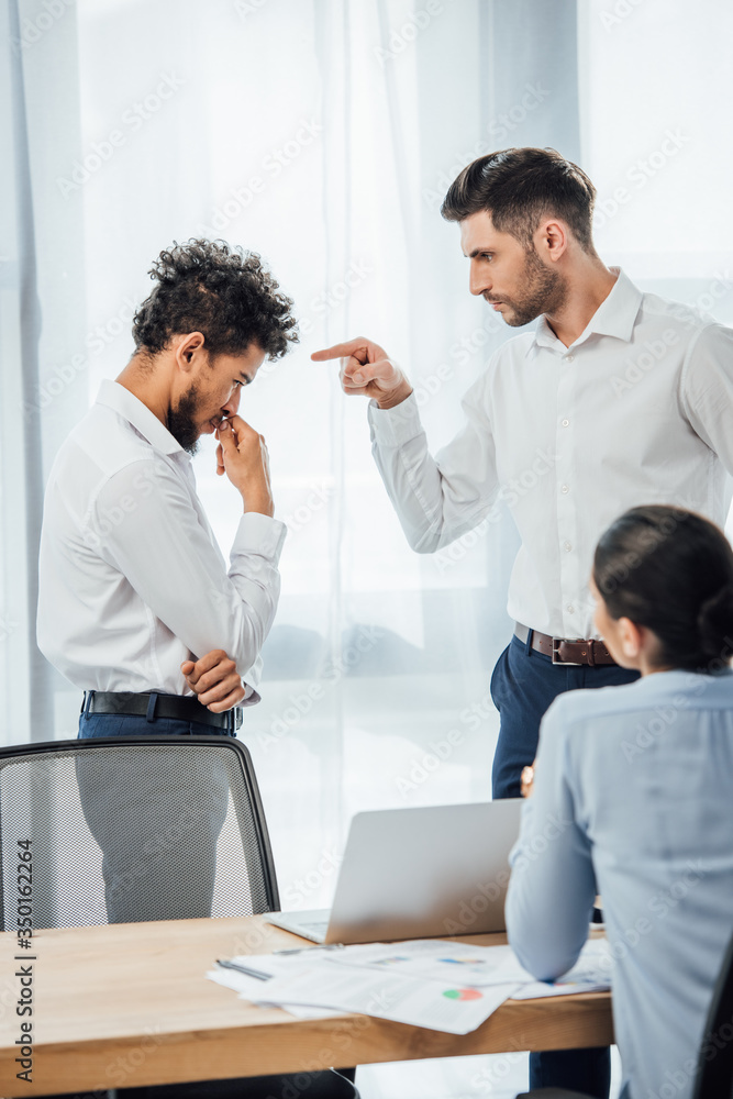 Selective focus of businessman pointing at offended african american colleague in office Selective focus of businessman pointing at offended african american colleague in office