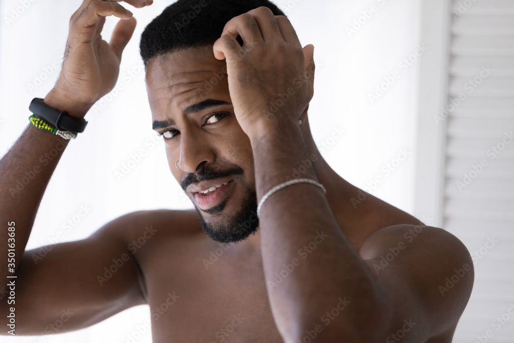 Naked millennial african American man look in mirror in bathroom style haircut  after shower in morning, handsome young biracial male get ready in home  bath, take care of hair, haircare concept Stock