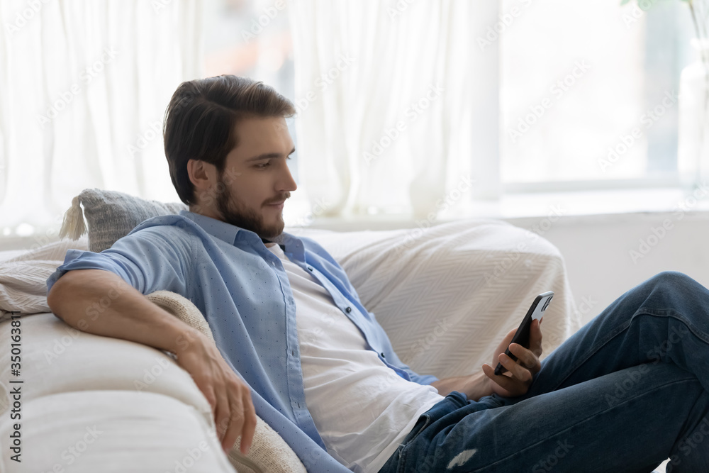 Young Caucasian man sit relax on sofa at home use modern smartphone gadget, relaxed millennial male rest on comfortable couch in living room browse wireless internet on cellphone, technology concept