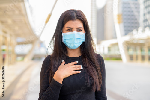 Sick young Indian woman with mask having sore throat in the city outdoors