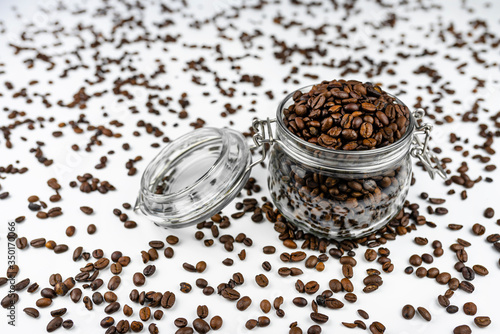 Fresh, roasted coffee beans in a open, transparent jar standing on a white table with lots of coffee scattered around.