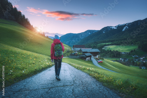 Beautiful young woman with backpack is standing on the mountain road at sunset in autumn. Landscape with sporty girl, rural road, green meadow, trees, purple sky in Italy at dusk in fall. Travel