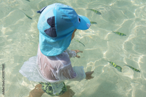 little boy in blue summer hat is trying to catch little striped bright fish in turquoise transparent sea, vacation activities for child