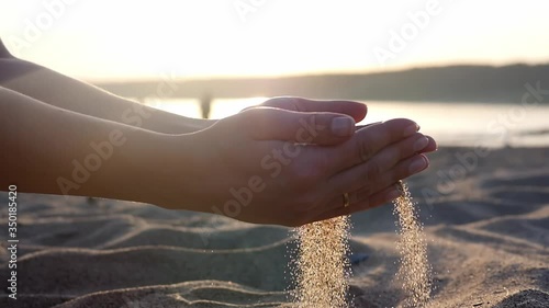 Close up of woman pouring sand running through fingers slow motion at the beach with sun flare and blue sky. Beautiful scene of young lady playing with sand on sunrise or sunset. Sand slowly pouring