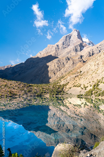The beautiful mountain trekking road with clear blue sky and rocky hills and the view of Alaudin lake in Fann mountains in Tajikistan © Aleksey