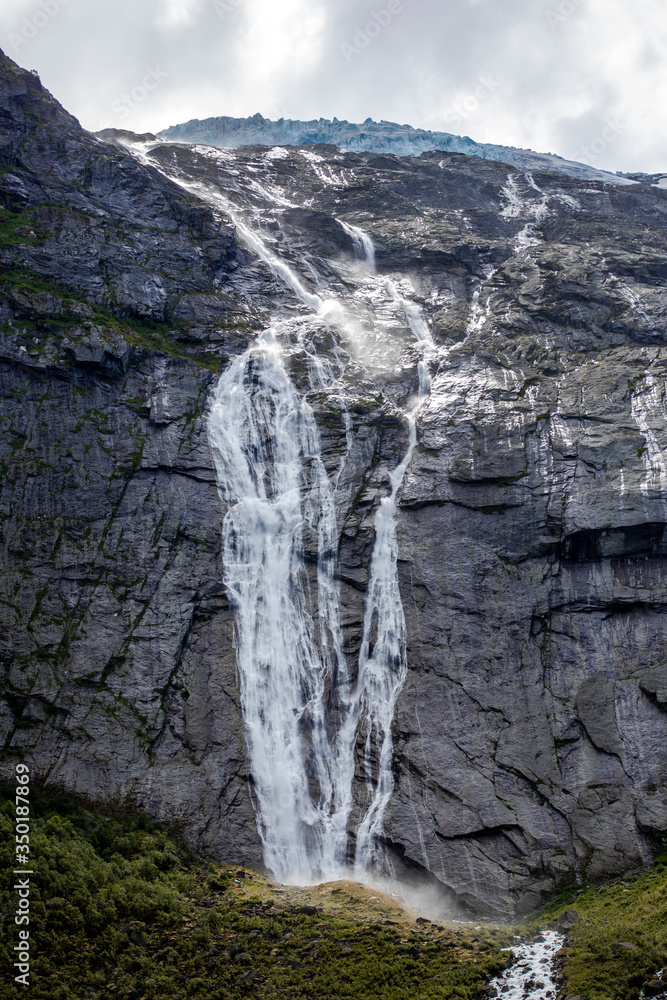 Melting of the Briksdalsbreen glacier with high waterfall from the giant rock mountain into the green valley on background of cloud sky. Vertical wallpaper. Norway