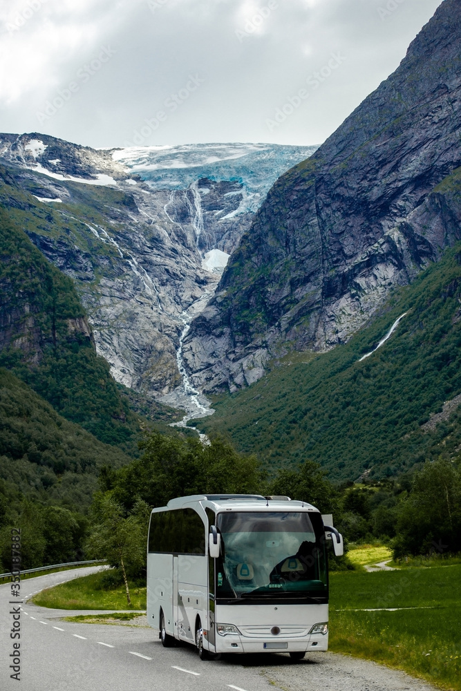 Big white tourist bus rides on the road. Blue ice tongue of Jostedal glacier melts from the giant rock mountains into the green valley with waterfalls. Vertical wallpaper. Norway