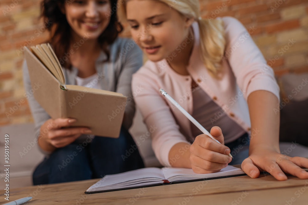 Selective focus of smiling woman holding book near child writing on notebook at home Selective focus of smiling woman holding book near child writing on notebook at home
