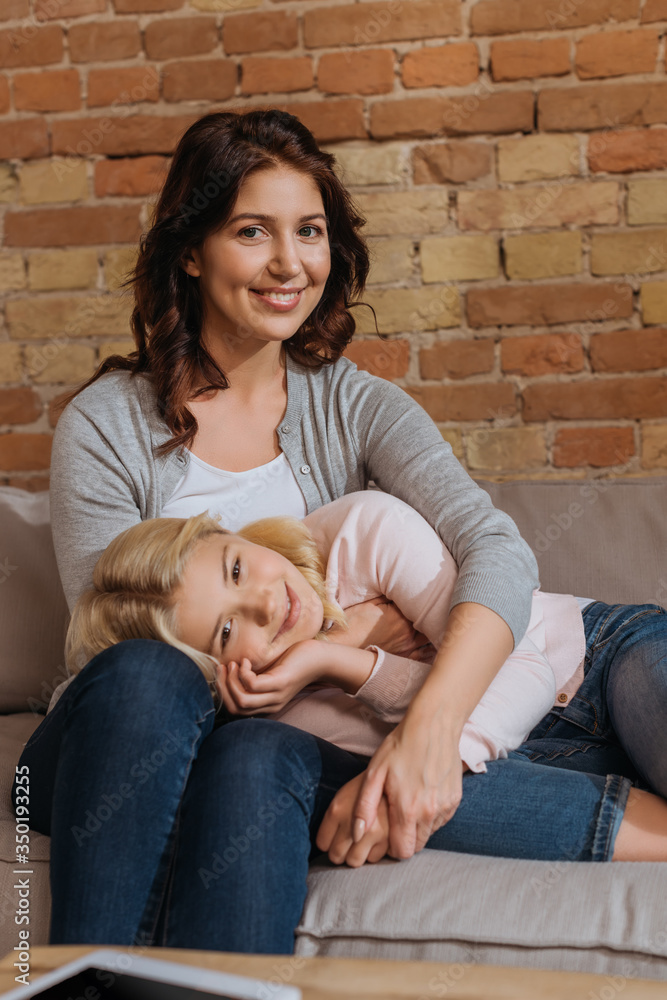 Selective focus of smiling mother and daughter looking at camera on sofa at home