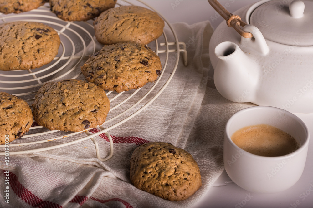 Several fresh cookies on a metal stand accompanied by a coffee maker and a white cup of coffee. Horizontal format.