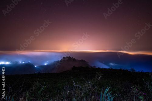 Mountains in fog at beautiful night in autumn in Dalat city, Vietnam. Landscape with Langbiang mountain valley, low clouds, forest, colorful sky with stars, city illumination at dusk.