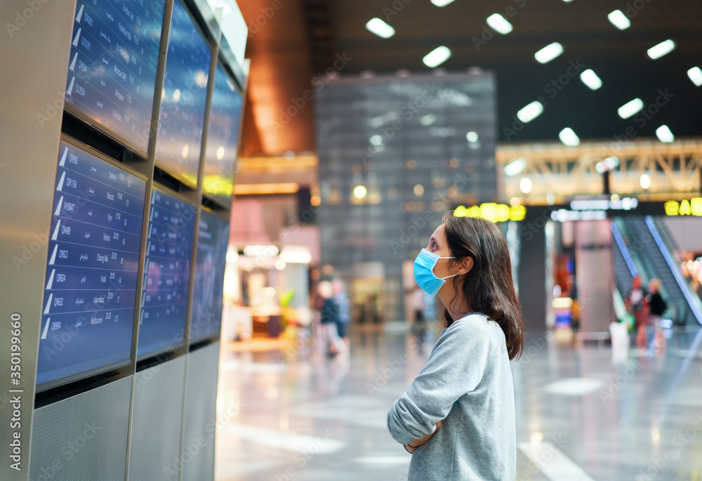 Woman in virus protection face mask looking at information board checking her flight in international airport - obrazy, fototapety, plakaty 