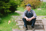 Senior gardener sitting on the steps in garden. Aged worker in overall and baseball cap resting after work. Portrait of old farmer