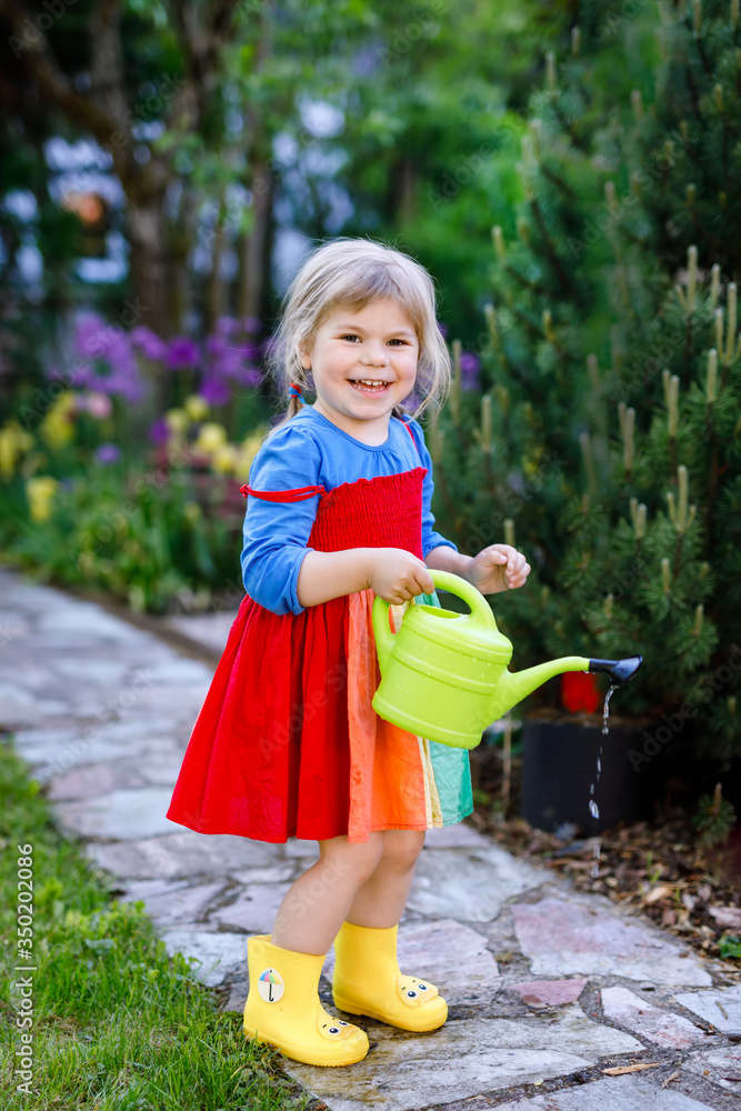 Φωτογραφία Beautiful little toddler girl in yellow rubber boots and colorful dress watering spring flowers with kids water can