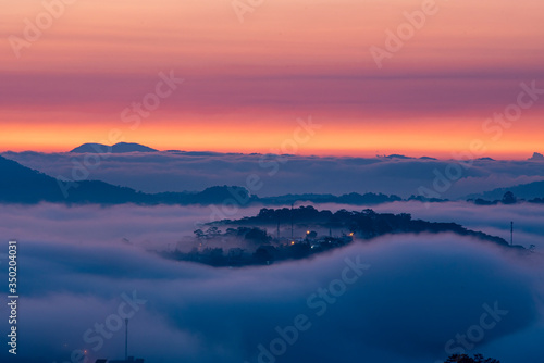 Mountains in fog at beautiful morning in autumn. Landscape with Langbiang mountain valley  low clouds  forest  colorful sky   city illumination at dusk.
